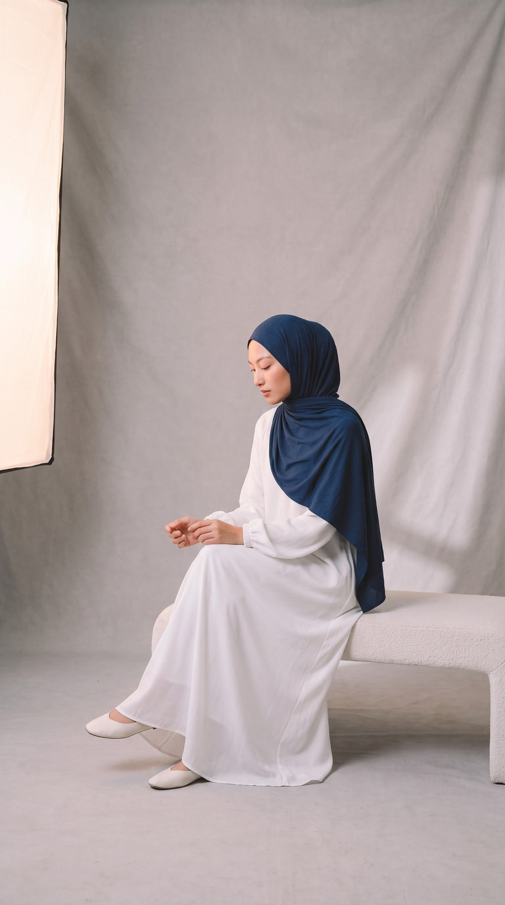 Eternal Aura Model trägt ein weißes langes Kleid mit einem dunkelblauen Hijab, sitzend auf einer Bank im Studio.